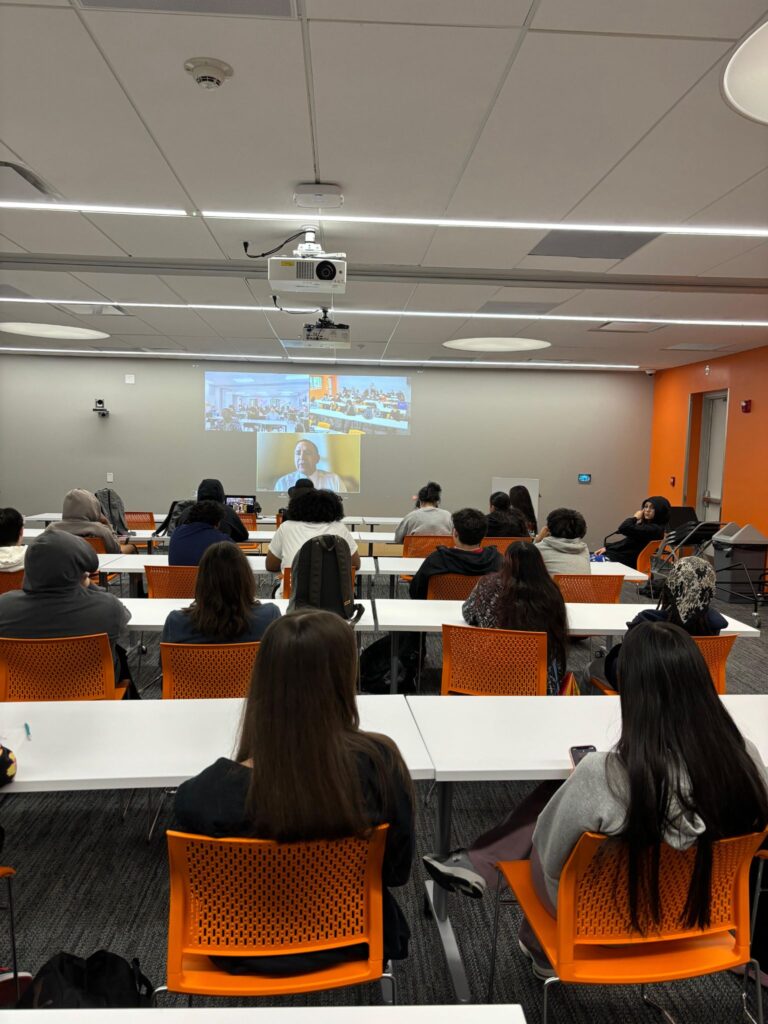 photograph taken from the back of a classroom facing a projection at the front of the classroom set up auditorium style with white desks and orange chairs. Seats filled with high school aged students